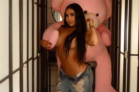 Brunette woman in denim jeans posing playfully with a giant pink teddy bear in a modern indoor setting.