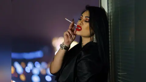 DeeaAnderson smokes a cigarette while wearing a black silk top and red lipstick against a dark background.