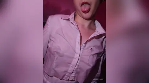 ChristyWeb sticks out her tongue while wearing a white button-down shirt against a pink background.