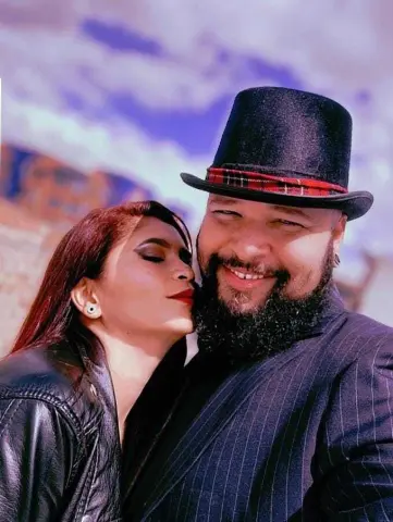 Smiling man in a black top hat and pinstripe suit embracing a woman in a black leather jacket under a blue sky.