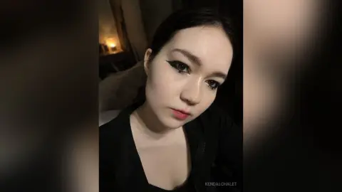 Young woman with winged eyeliner and black outfit in a moody, dimly lit indoor setting.