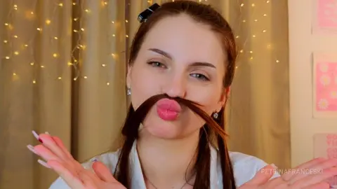 Playful young woman with long brown hair making a funny mustache face in a cozy room with fairy lights.