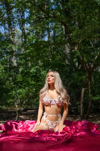 Blonde woman in white lace lingerie sitting on pink satin fabric in a lush green forest setting.
