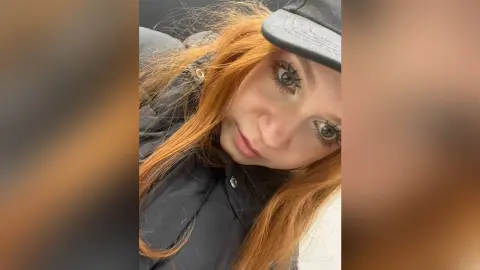 Redheaded woman in a black jacket and baseball cap looking thoughtfully into the camera indoors.