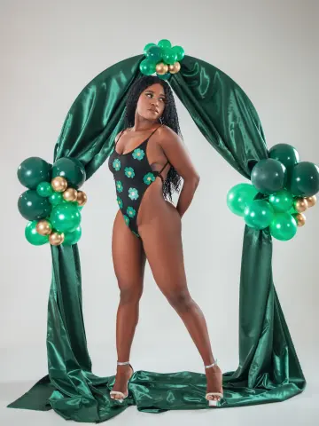 Curvy woman in a black floral swimsuit posing against a green satin drape and balloon backdrop.