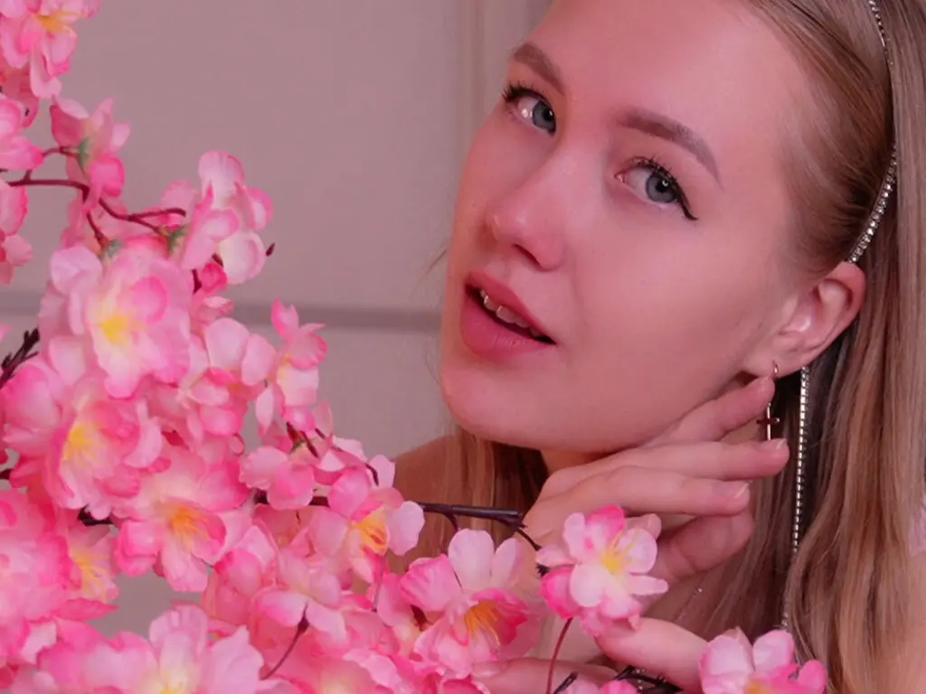 Blonde woman with soft makeup posing near pink cherry blossoms in a dreamy, romantic setting.