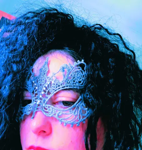 Woman with curly dark hair wearing a silver masquerade mask in moody blue lighting.