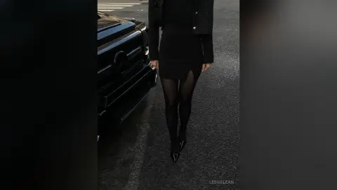 Elegant woman in a chic black mini dress and tights standing next to a luxury car on a city street.