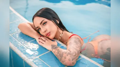 Tattooed woman in a red bikini relaxing by a bright blue swimming pool on a sunny summer day.