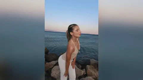 AmberHazel poses by the ocean at sunset, wearing a white crochet halter top and white pants with her hair in a ponytail.