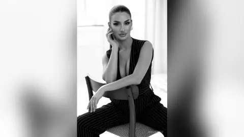 AnnaKarev sits on a chair wearing a low-cut black pinstripe jumpsuit with her hair pulled back in a sleek ponytail.