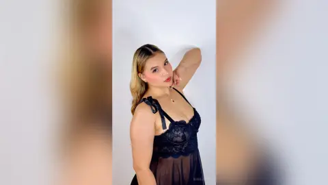 Blonde woman in a black lace camisole posing against a white wall with a soft, elegant mood.