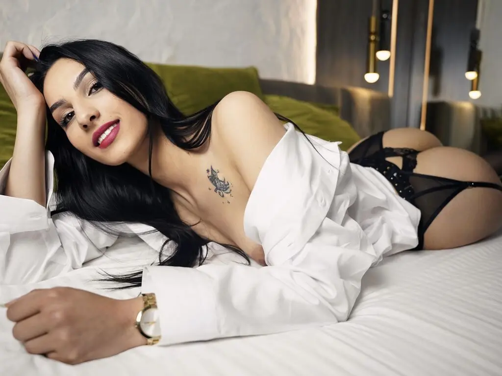 Beautiful brunette woman in a white oversized shirt relaxing on a bed in a cozy, intimate bedroom setting.