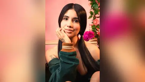 Smiling young woman in a dark green off-the-shoulder top posing against a pink floral background.