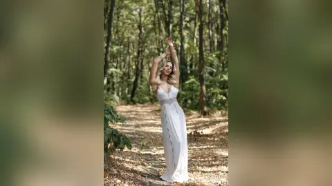 Blonde woman in a flowing white maxi dress posing gracefully on a sunlit forest path in a dreamy woodland setting.