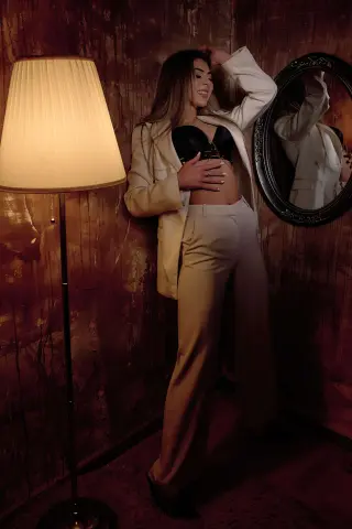 Elegant woman in a white suit and black bra posing in a moody, dimly lit room by a floor lamp.