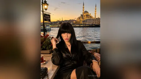 SofiaBleir — SofiaBleir sits at an outdoor cafe table by the water at sunset wearing a black satin dress.