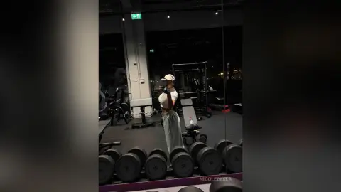 NicoleBella stands in a gym wearing a white crop top and light grey sweatpants while taking a mirror selfie.