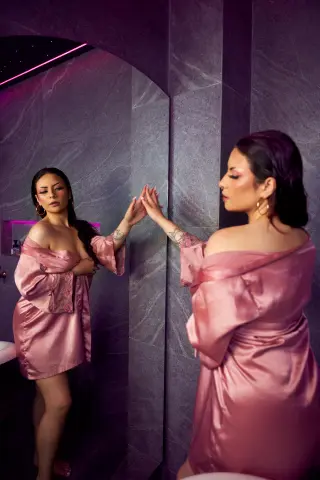 Elegant woman in a pink satin robe gazing into a mirror in a moody, dimly lit modern bathroom.