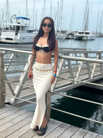 Woman in a white bikini top and long skirt posing on a wooden pier near sailboats under a bright sky.
