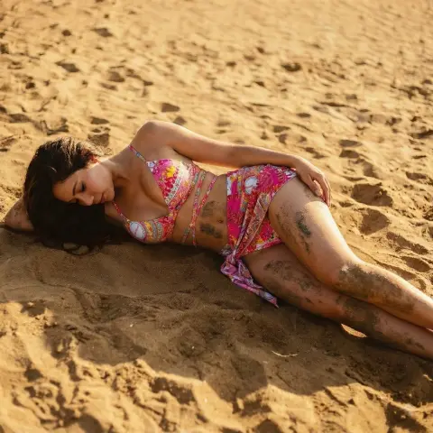 LilithVelluto69 lies on the sand wearing a pink patterned bikini, showing her tanned skin and long dark hair.