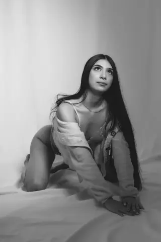 Moody black and white portrait of a long-haired woman in a white shirt sitting on a draped fabric background.
