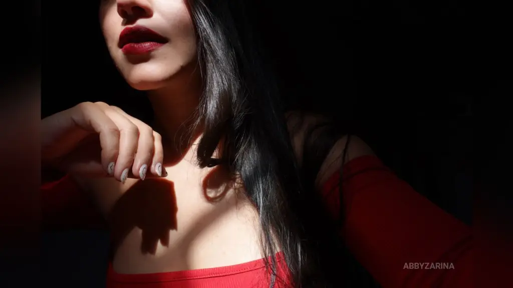 Nude woman with long dark hair and large rose tattoo standing by a bathroom sink in a bright modern room.