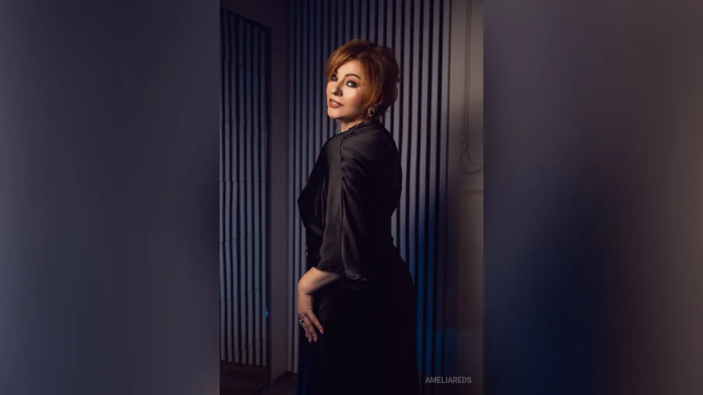 AmeliaReds — AmeliaReds poses in a black silk blouse and dark trousers against a blue slatted wall.