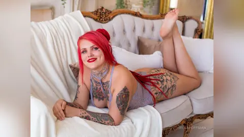 Smiling woman with red hair and tattoos relaxing on a white vintage sofa in a bright, cozy room.