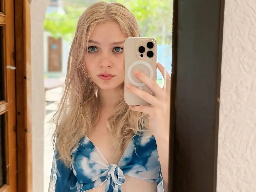 Blonde woman in a blue tie-dye crop top taking a mirror selfie in bright natural light.