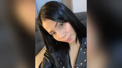 Smiling woman with long dark hair wearing a black leather jacket in a cozy indoor setting.