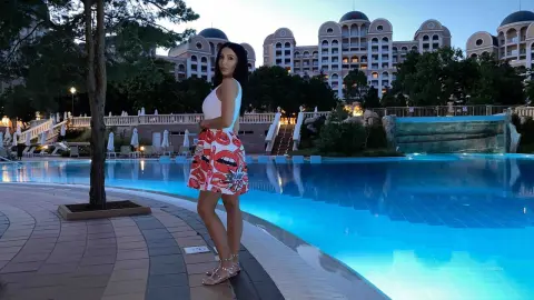 Woman in white tank top and red patterned skirt standing by a luxury resort swimming pool at twilight.