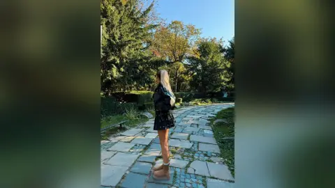 Blonde woman in a black outfit walking on a stone path through a lush green summer garden.