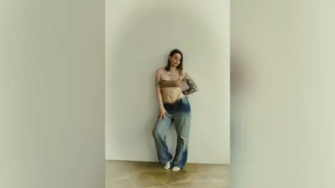 Young woman in a tan crop top and baggy blue jeans posing against a minimalist white wall.