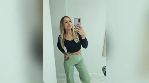 Jess poses in a mirror wearing a black long-sleeve crop top and light green trousers, showing off her blonde hair and slim waist.
