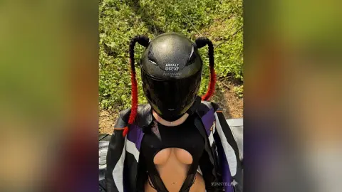 Woman in black bodysuit and leather jacket wearing a black motorcycle helmet in a lush green outdoor setting.