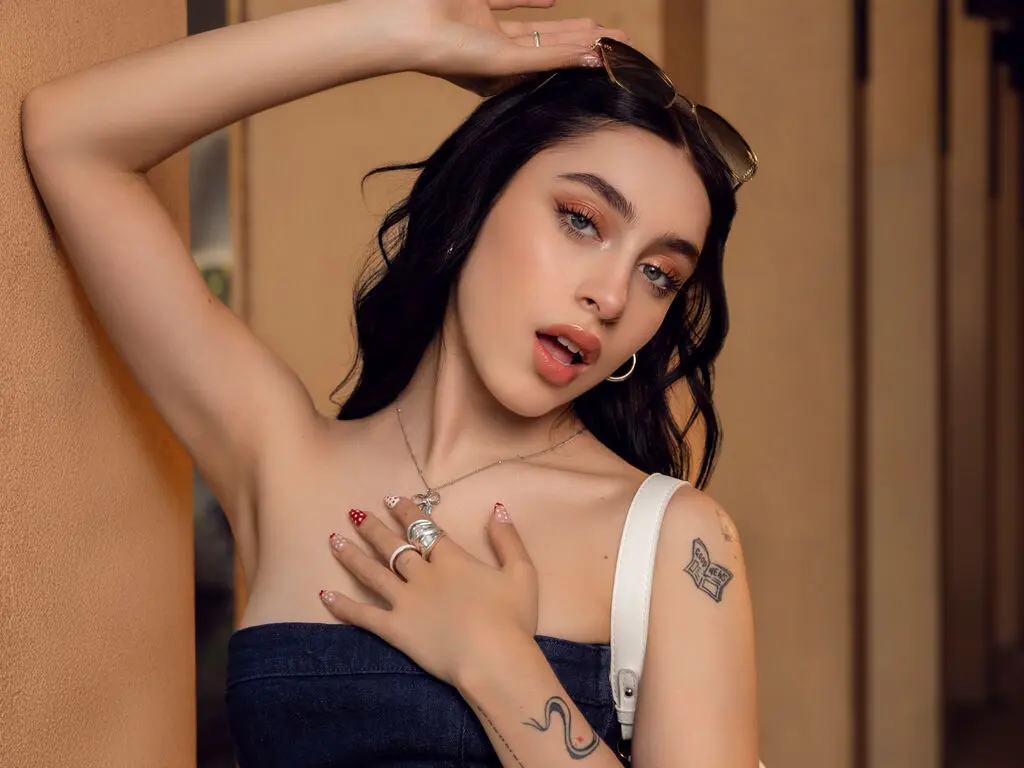 Young woman with dark hair and tattoos wearing a blue tube top and silver jewelry in a warm indoor setting.