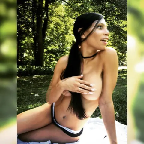 Young woman in black bikini bottoms relaxing on a white towel in a sunny, lush green forest setting.