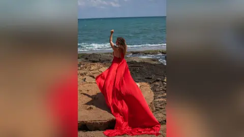 NataliaNacher stands on a rocky shore facing the ocean, wearing a long, flowing red dress with her arm raised against a blue sky.