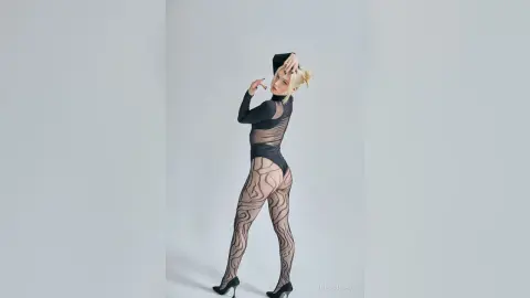 Blonde woman in a black sheer patterned bodysuit posing against a minimalist grey studio background.