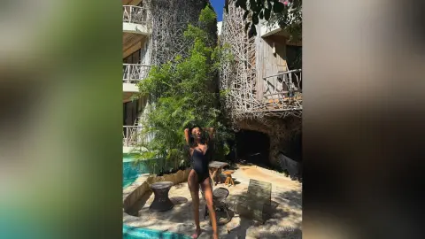 Woman in black one-piece swimsuit relaxing by a tropical poolside patio near lush green plants.