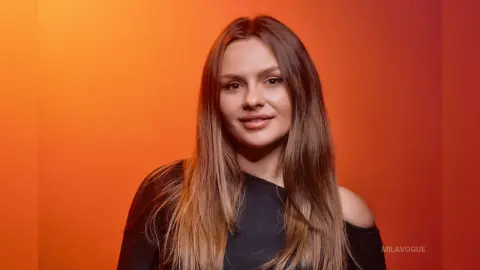 MilaVogue poses with long blonde-tipped hair, wearing a black off-the-shoulder top against a warm orange background.