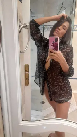 Young woman in a sheer black polka dot robe taking a mirror selfie in a bright bathroom.