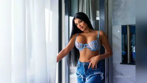 Beautiful woman with long dark hair in a light blue lace bra and denim shorts standing by a bright window.