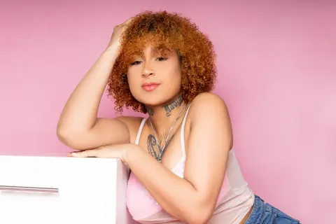 Young woman with curly hair and tattoos wearing a white tank top against a pink background.