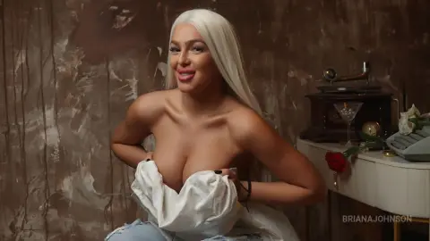 Smiling woman with long white hair in a white shirt sitting in a vintage room with a retro aesthetic.