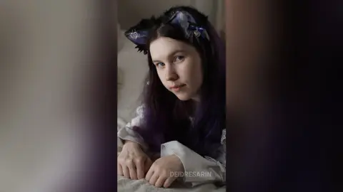 Young woman with purple hair wearing cat ear headband and white loungewear in a soft, moody indoor setting.