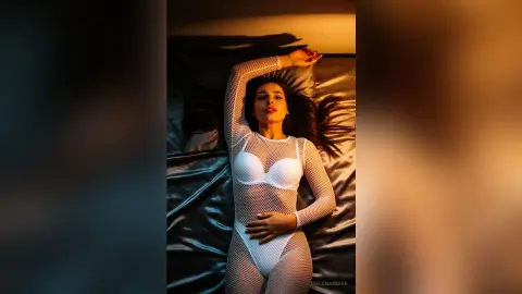 Woman in a white mesh bodysuit relaxing on a dark satin bed in moody, warm lighting.
