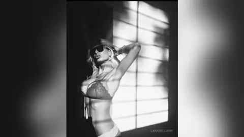 Moody black and white portrait of a blonde woman in lace lingerie posing by a sunlit window with shadows.
