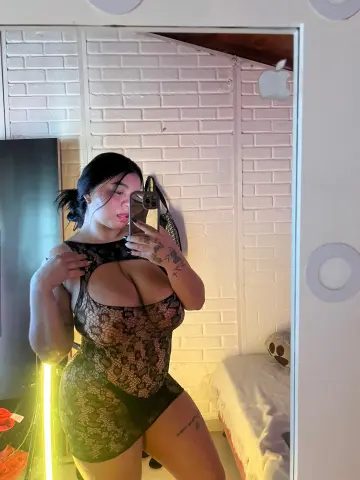 Young woman in a black lace bodysuit taking a mirror selfie in a bright room with white brick walls.